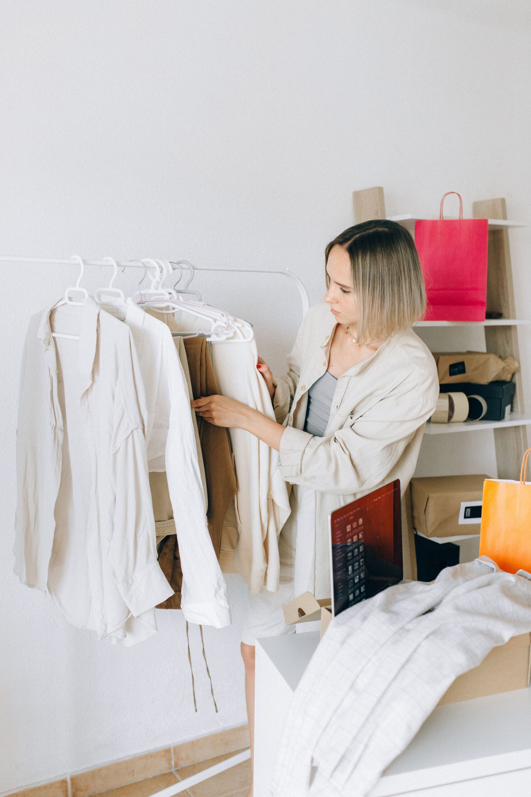 Woman organizing neutral-toned clothing on a garment rack in a minimalist fashion workspace with shopping bags and accessories.